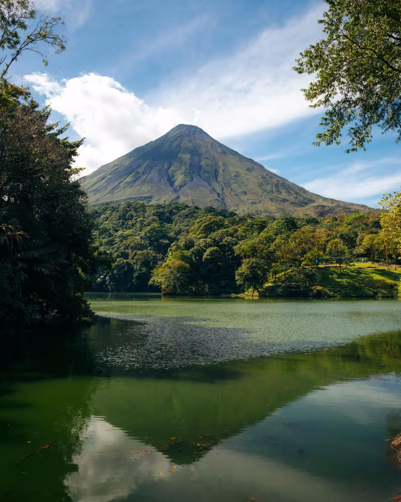 A conical volcano rises above a dense green forest and reflects in the calm water of a lake.